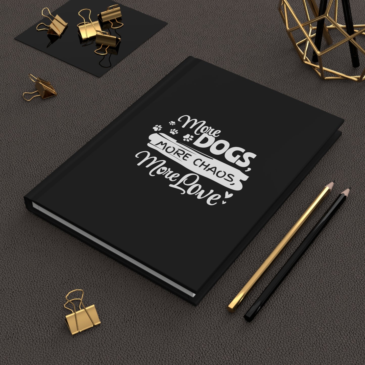 Black notebook with 'More Dogs, More Chaos, More Love' text on a dark surface with office supplies.