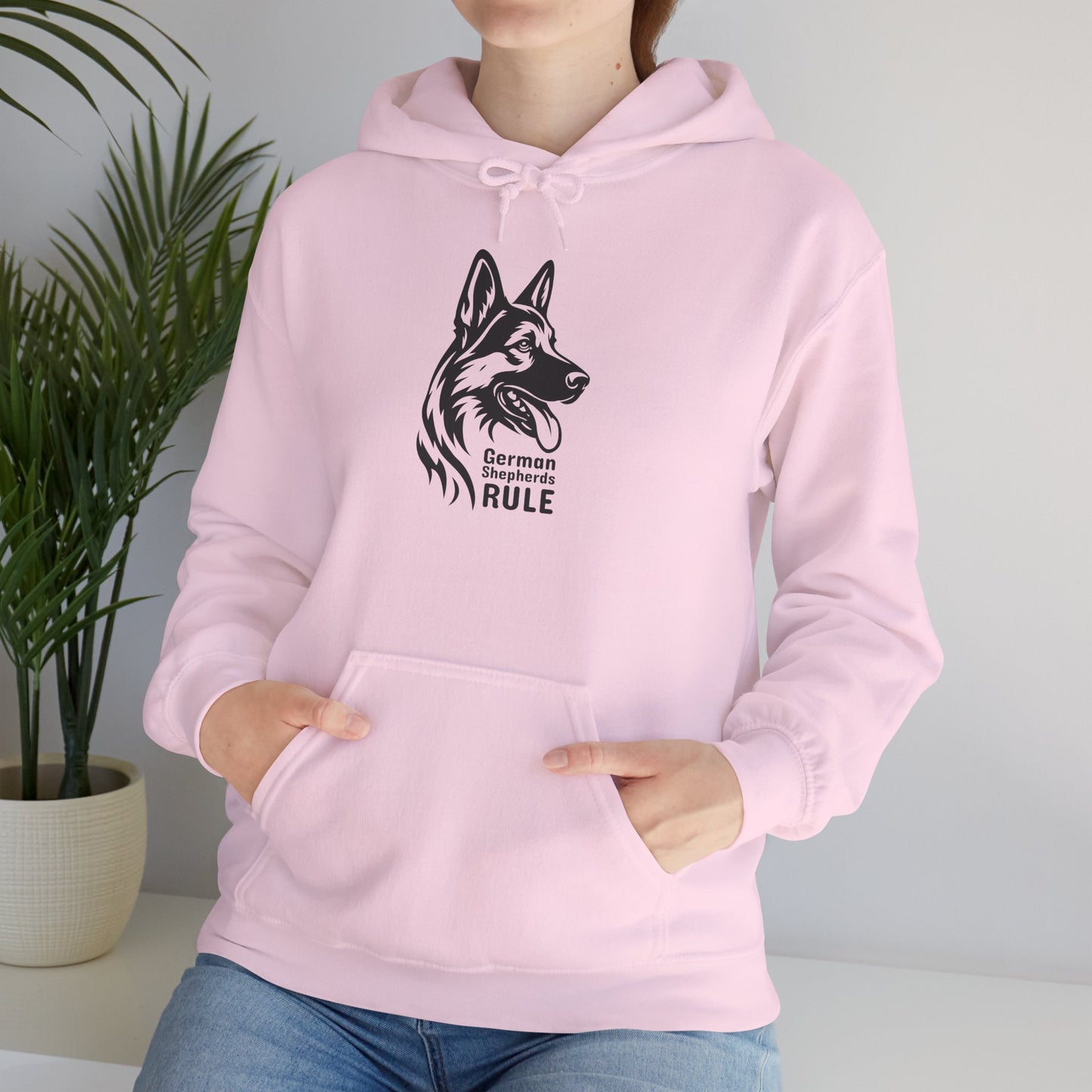 Person wearing a pink hoodie with a graphic of a German Shepherd and text, standing indoors next to a plant.