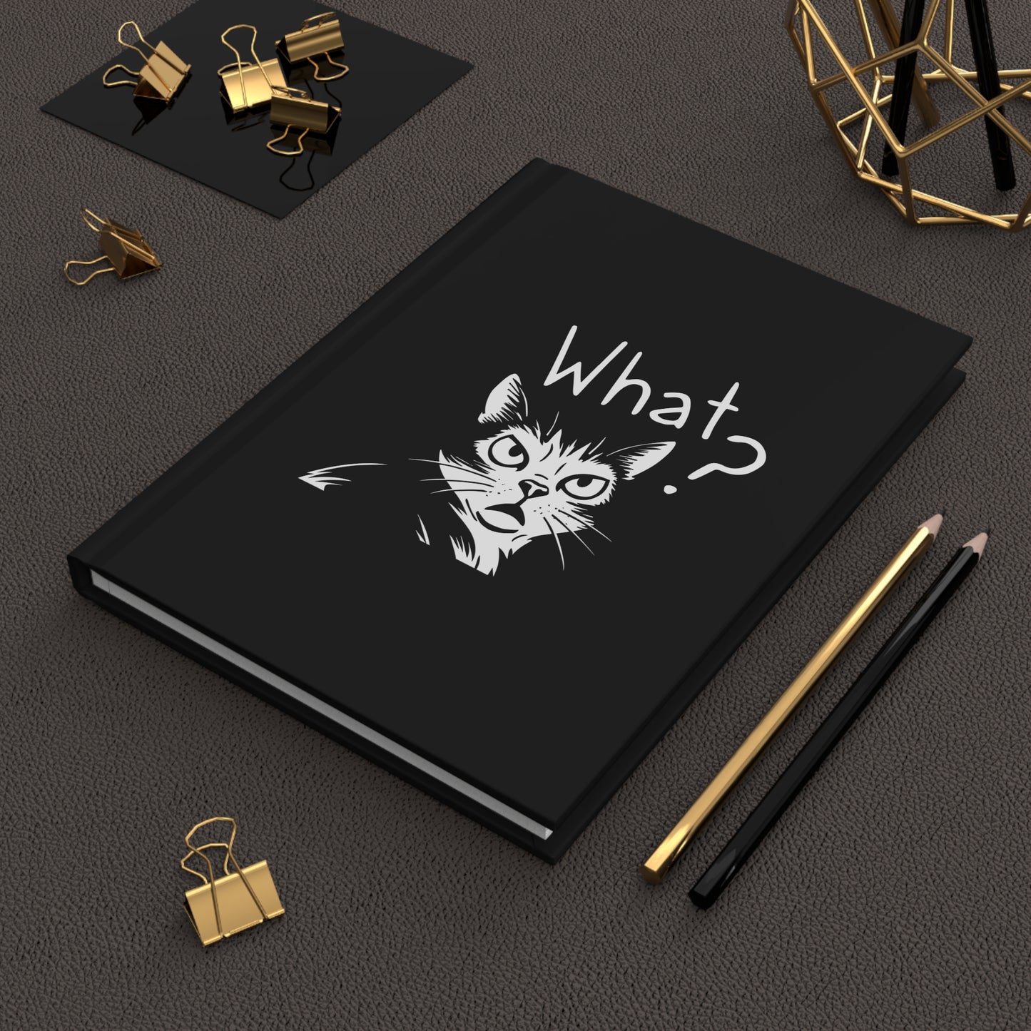 Black notebook with a cat illustration and 'What?' text on a dark surface with office supplies.