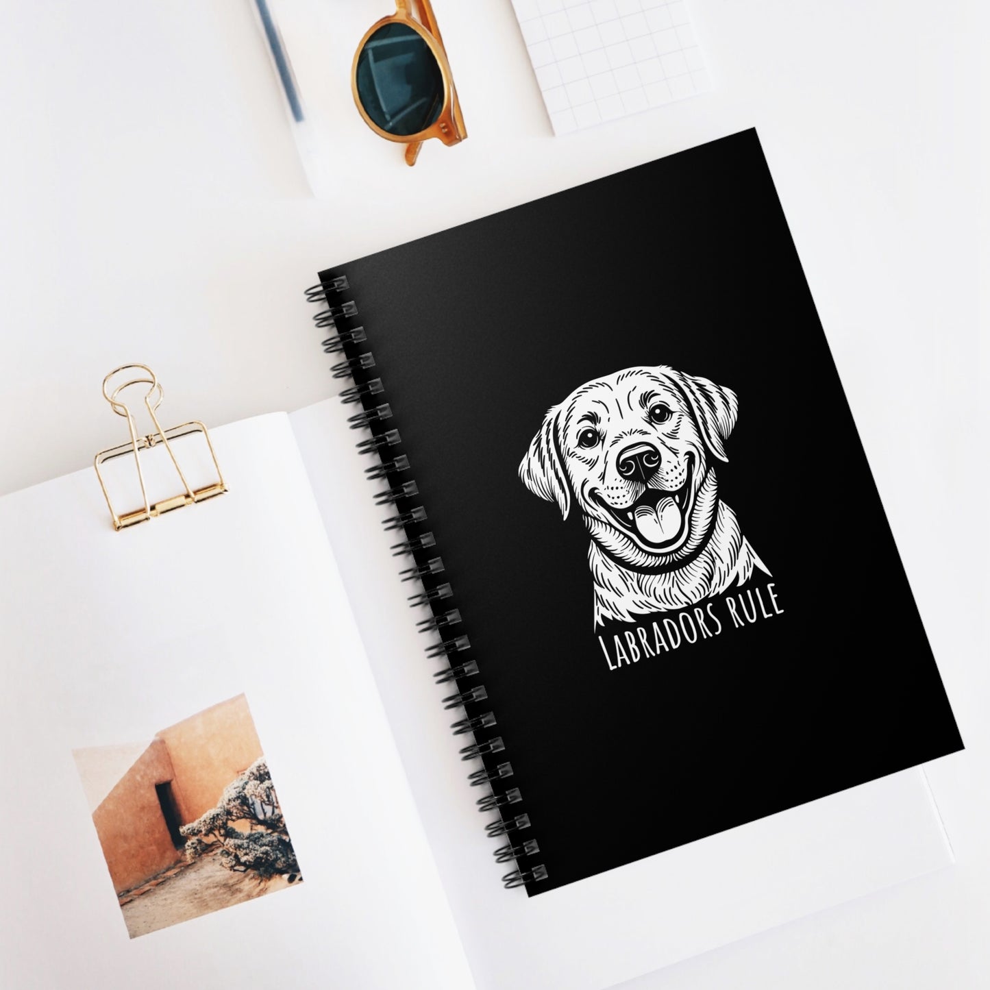 Black notebook with a dog illustration and 'Labradors Rule' text on a white surface with sunglasses and a photo clip.