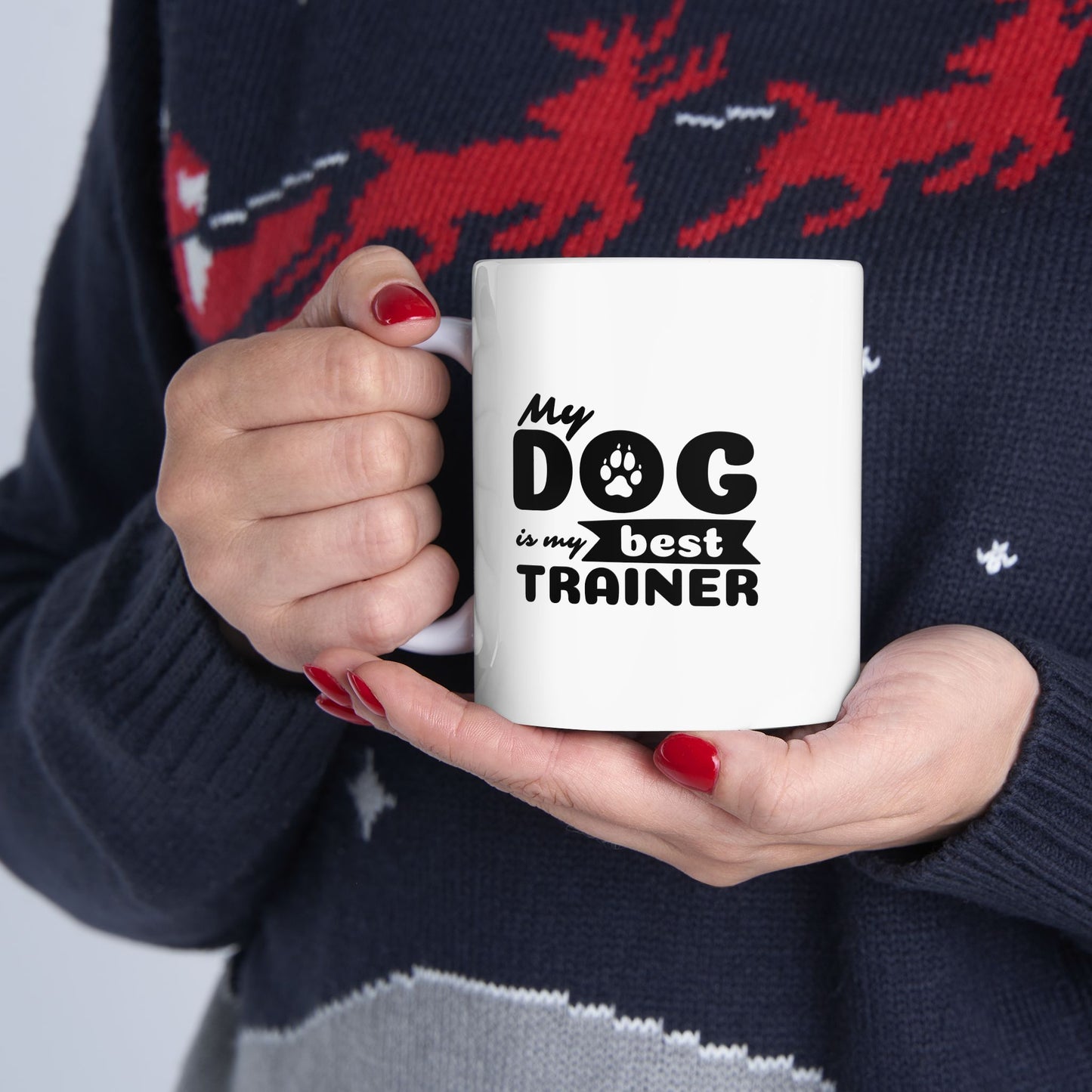 Person holding a white mug with 'My dog is my best trainer' text, wearing a dark sweater with red deer design.