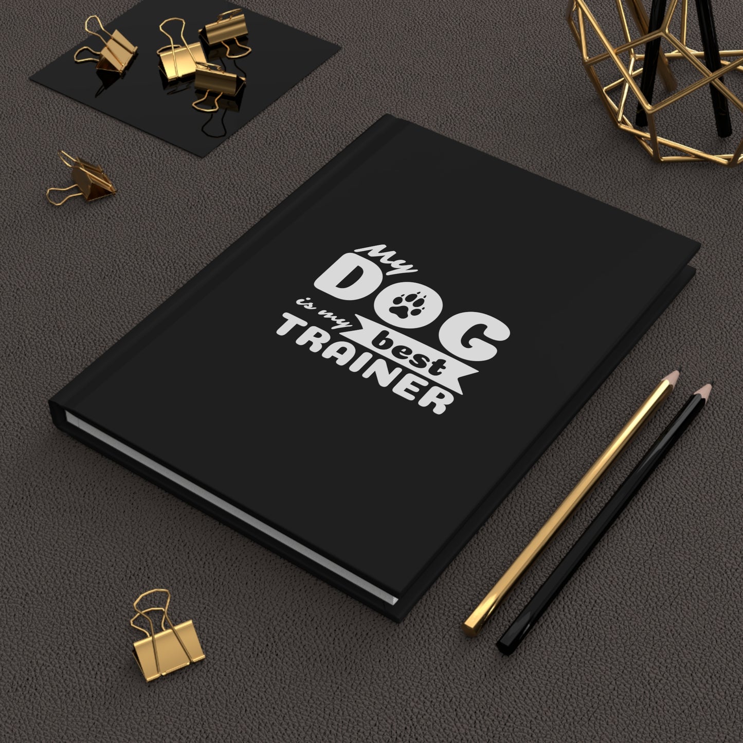 Black notebook with 'My dog is my best trainer' text on a dark surface with office supplies.