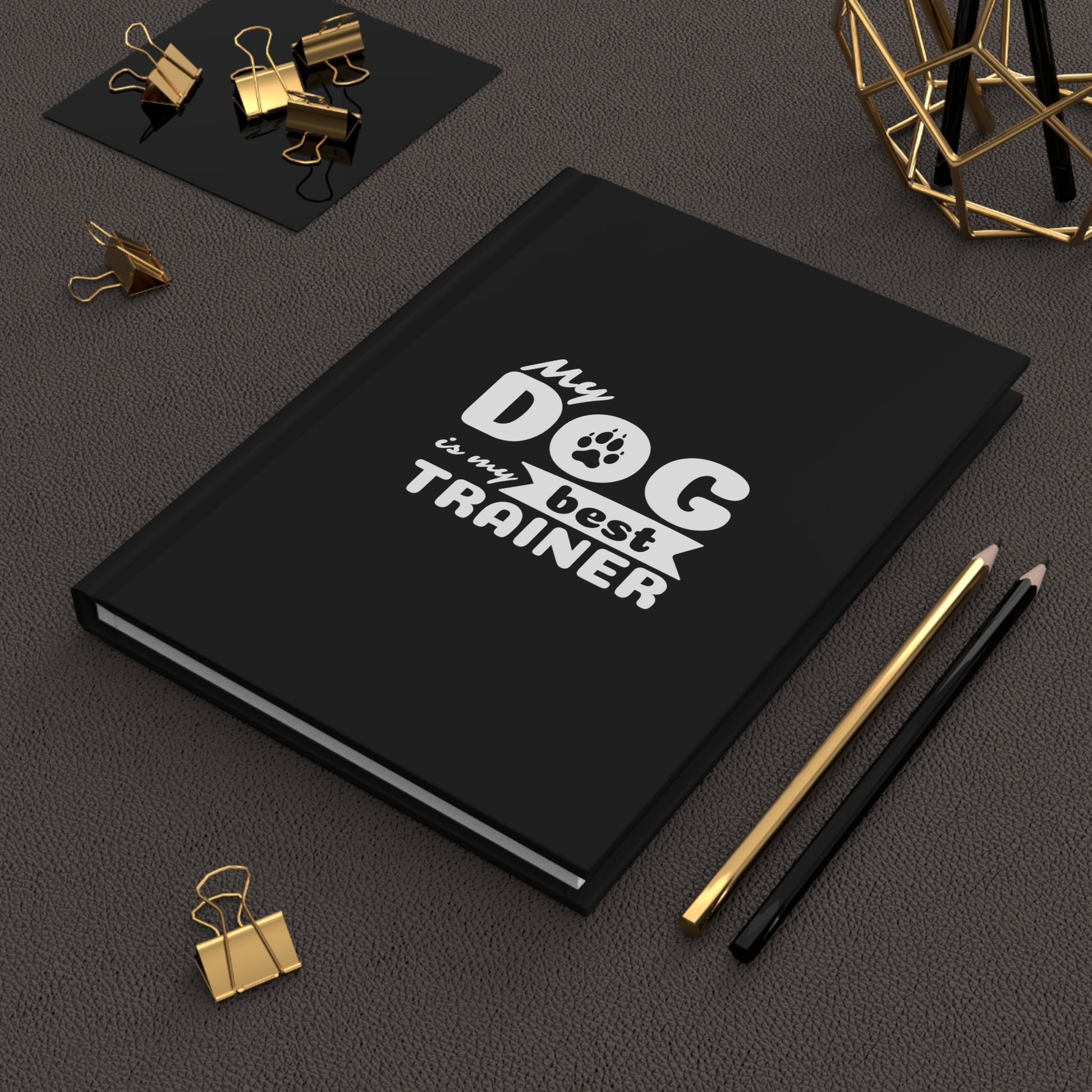 Black notebook with 'My dog is my best trainer' text on a dark surface with office supplies.