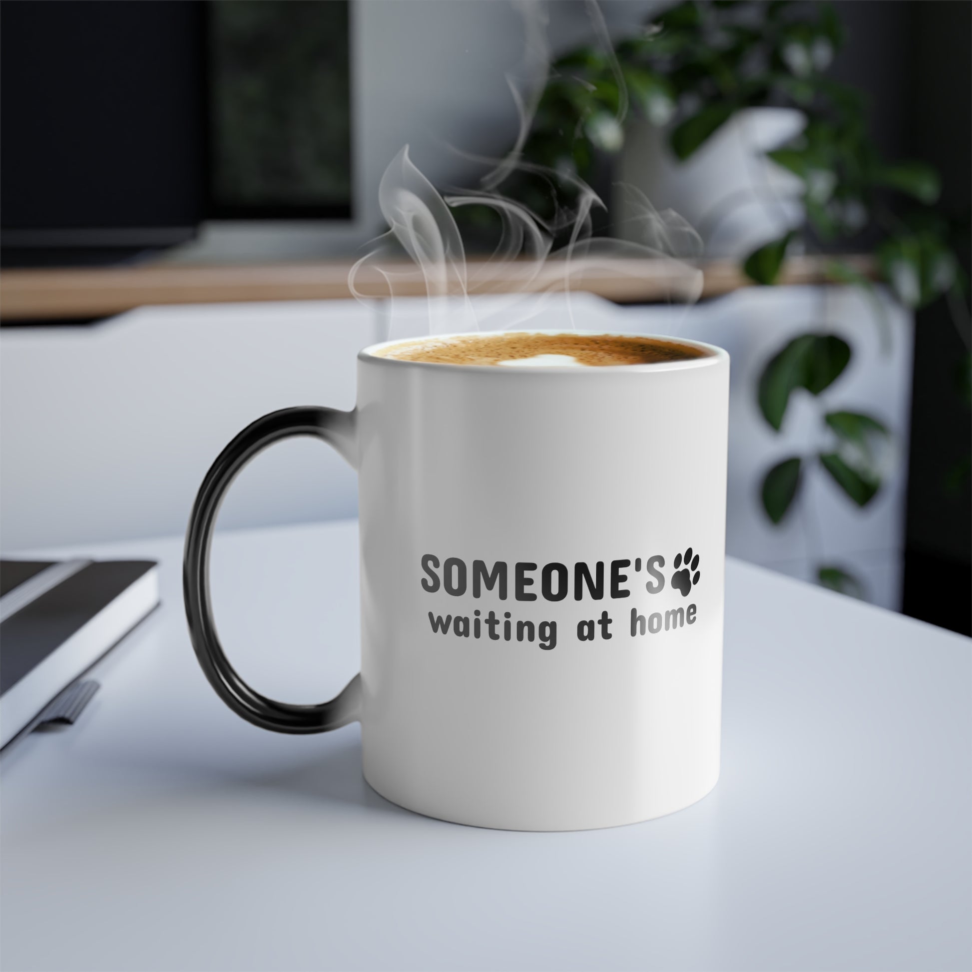 White mug with black handle and text 'SOMEONE'S waiting at home' on a desk with steam rising from it.