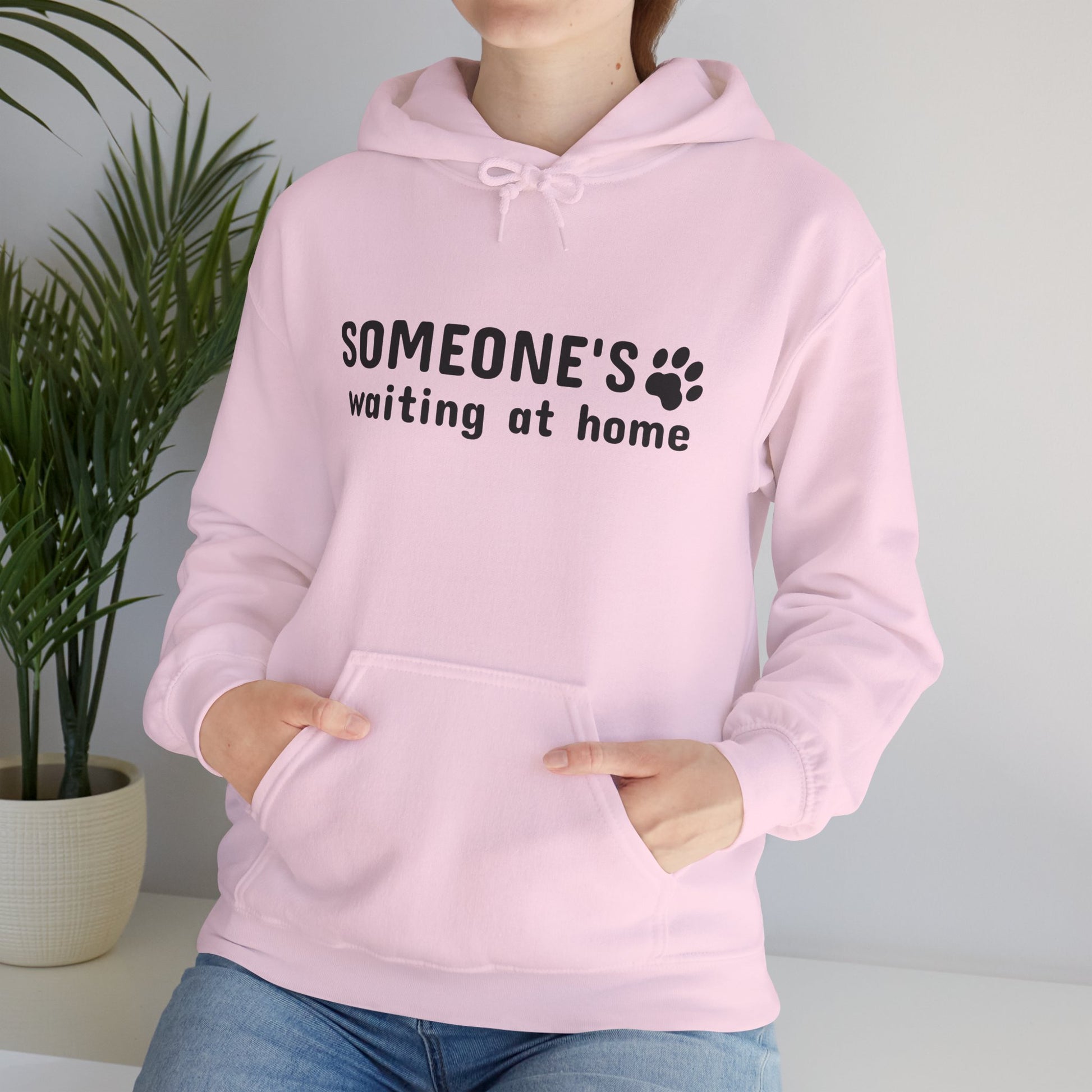 Person wearing a pink hoodie with text and paw print design, standing indoors next to a plant.