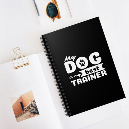 Black notebook with 'My dog is my best trainer' text on a white surface with sunglasses and photos.
