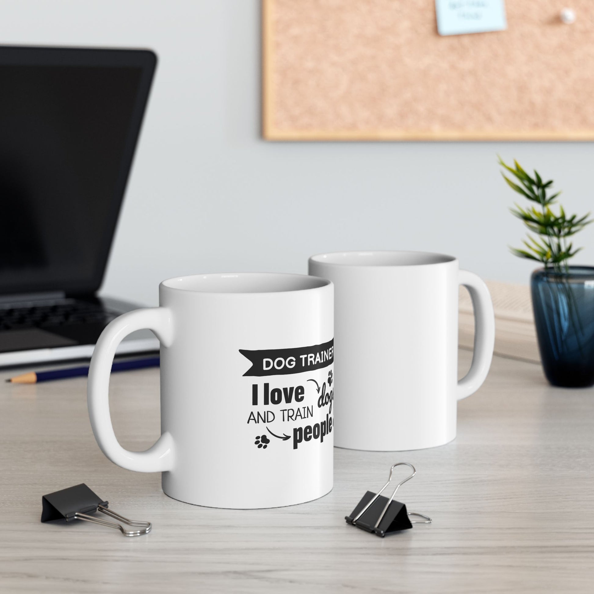 Two white mugs with text on a desk with office supplies and a plant.