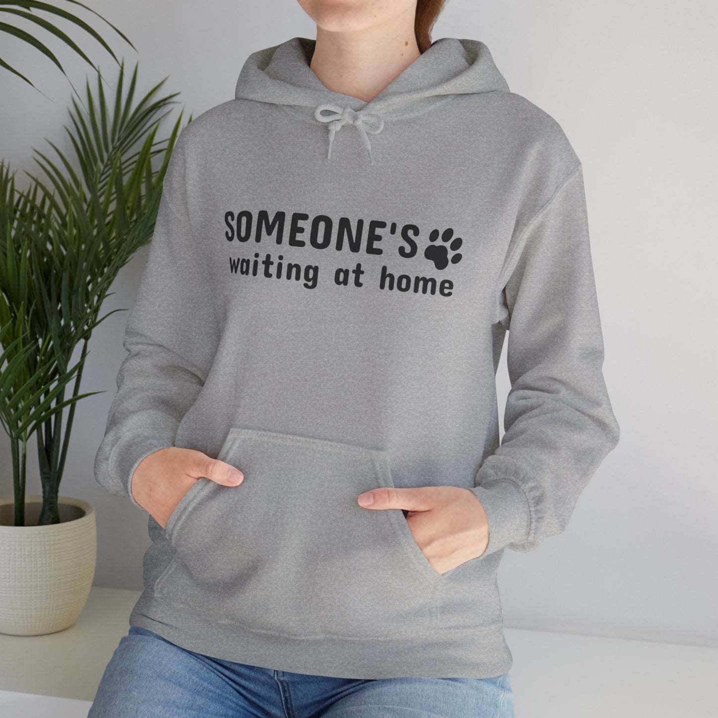 Person wearing a gray hoodie with text and paw print design, standing indoors with a plant in the background.
