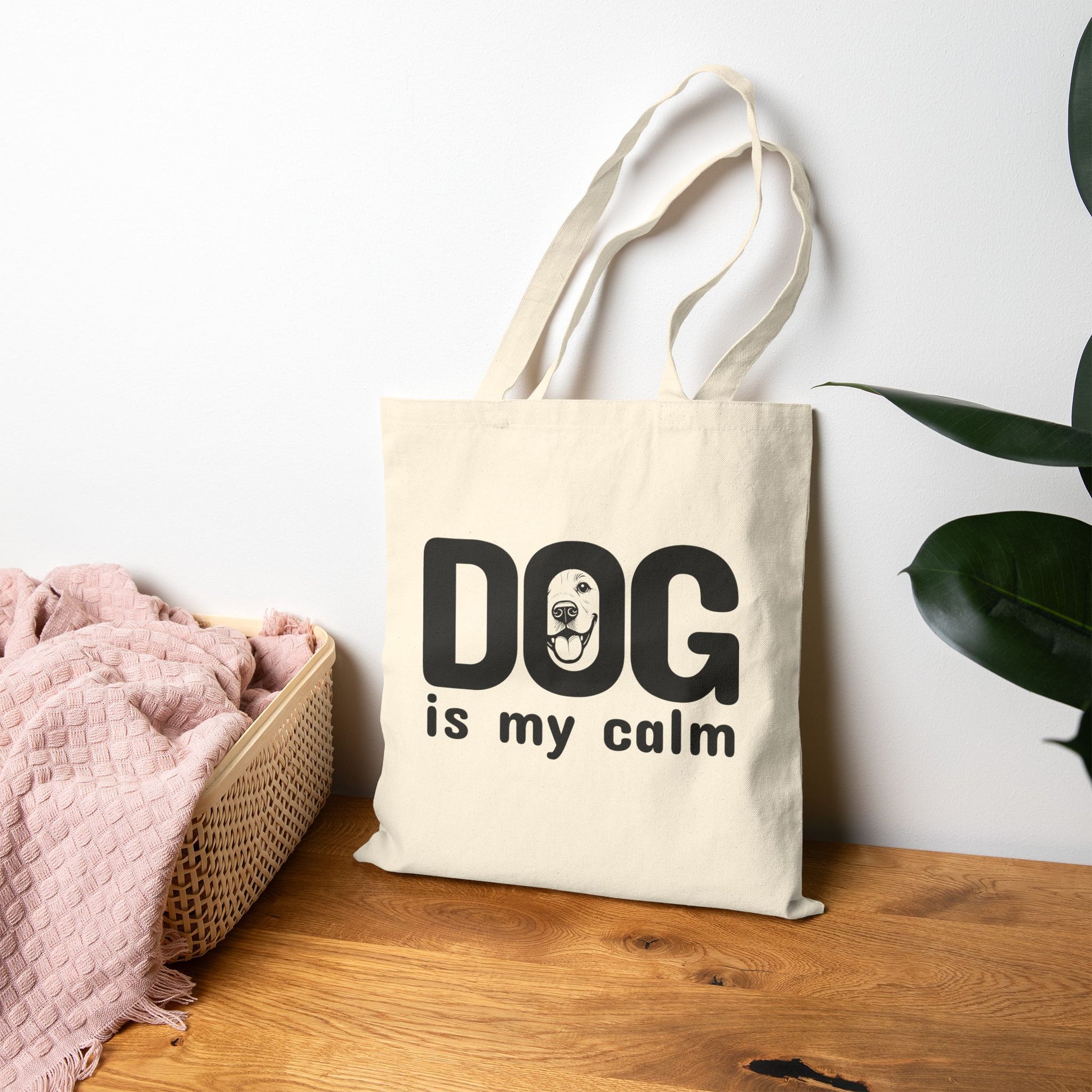 Beige tote bag with 'DOG is my calm' text on a wooden surface with a white wall and plant in the background