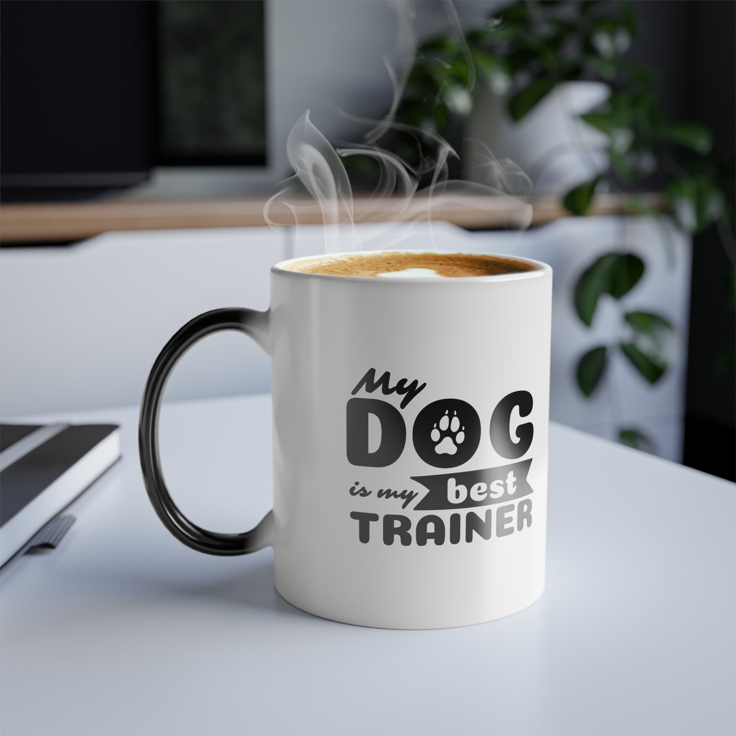 White mug with black handle and text 'My dog is my best trainer' on a desk.
