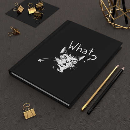 Black notebook with a cat illustration and 'What?' text on a dark surface with office supplies.