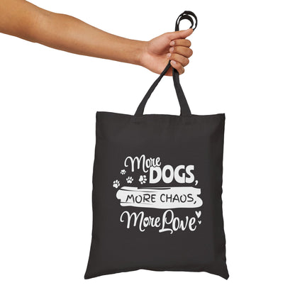 Black tote bag with white text 'More Dogs, More Chaos, More Love' held by a hand on a white background