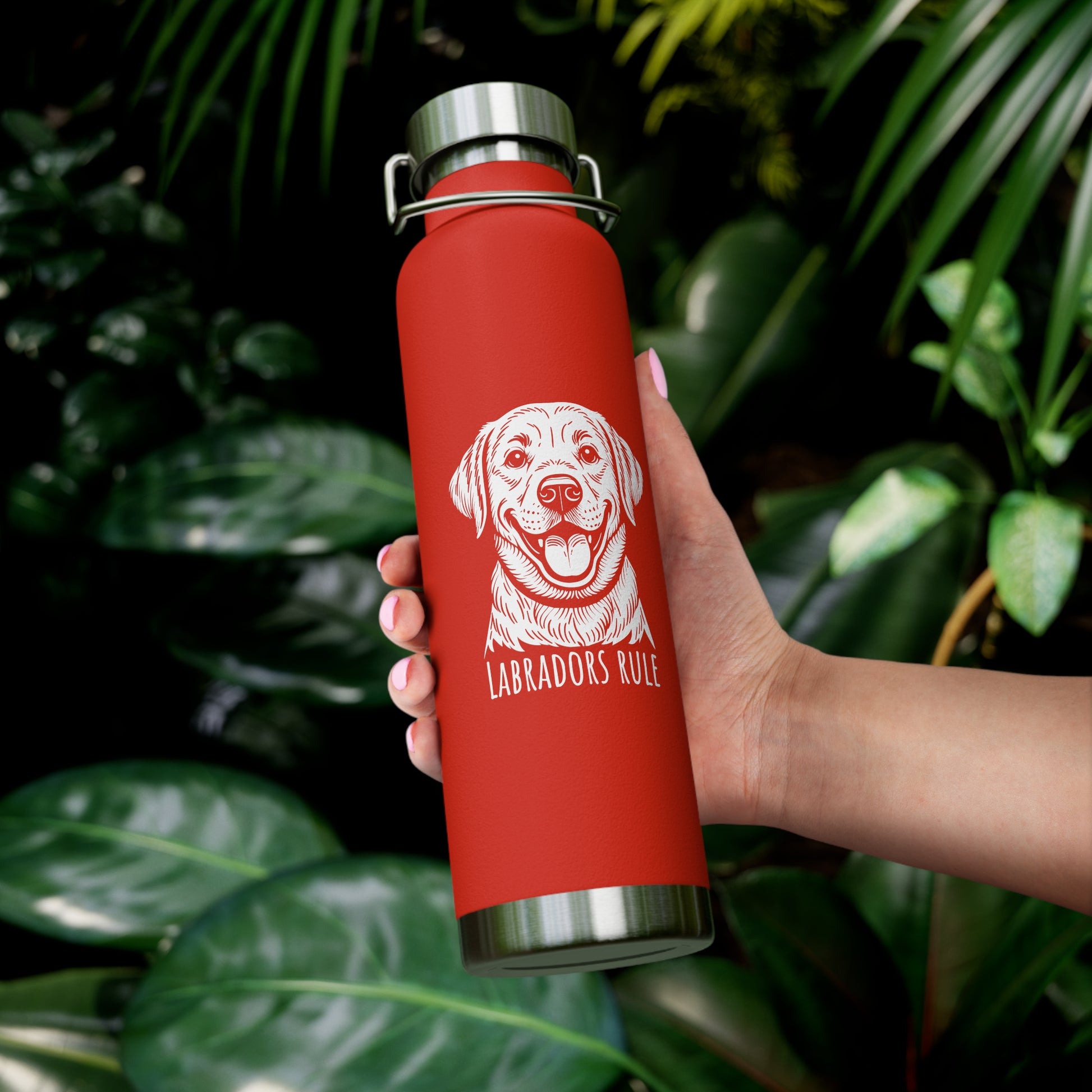 Red water bottle with a dog illustration and 'Labradors Rule' text held by a hand against a leafy background