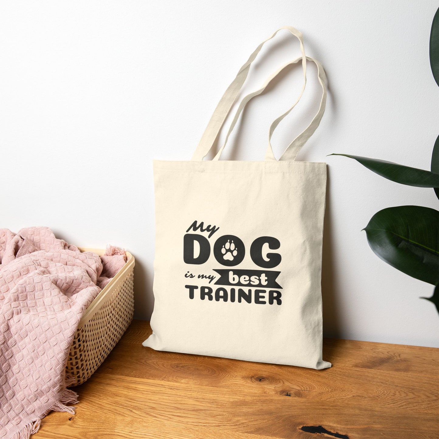 Tote bag with 'My dog is my best trainer' text on a wooden surface with a white wall and plant in the background
