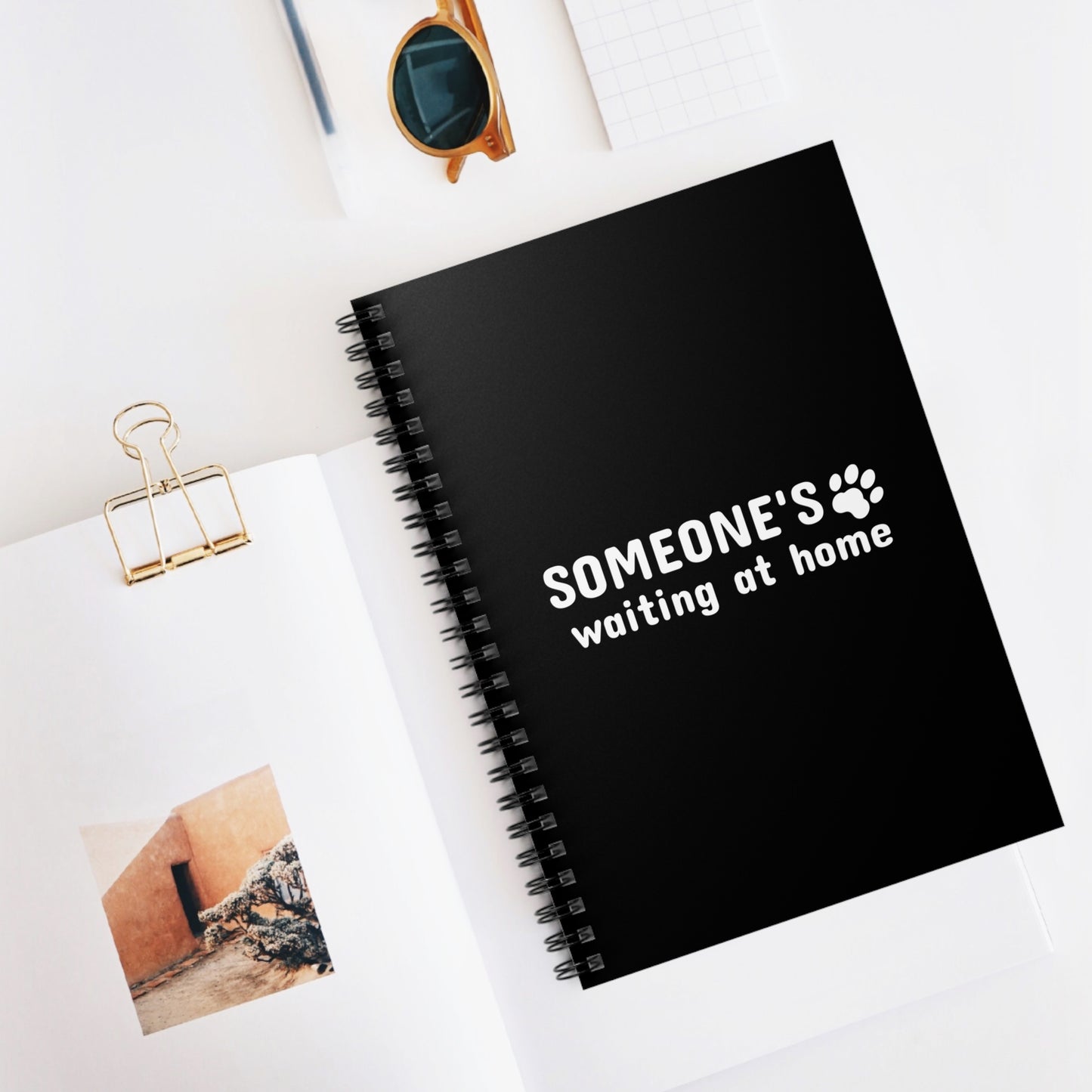 Black notebook with 'SOMEONE'S waiting at home' text on a white surface with a photo and sunglasses.