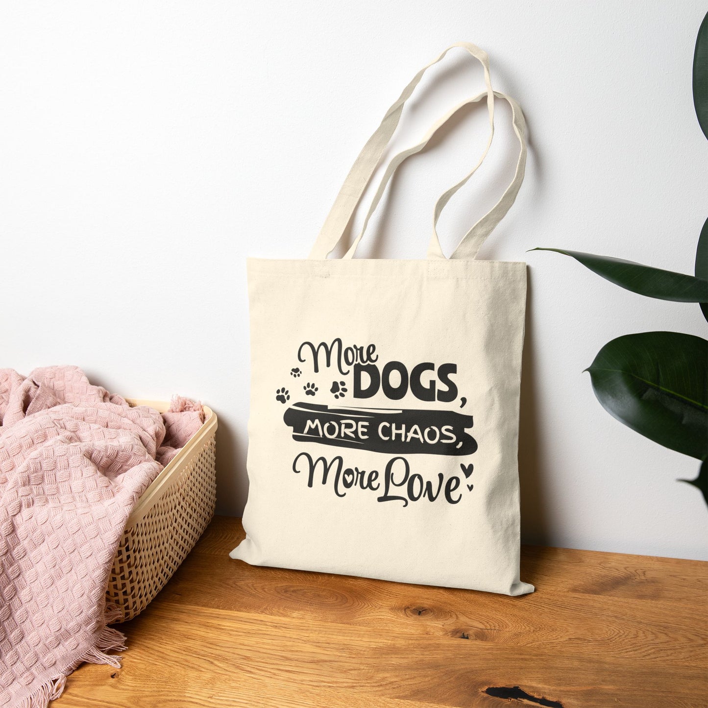 Tote bag with 'More Dogs, More Chaos, More Love' text on a wooden surface with a plant in the background