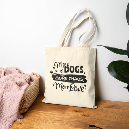 Tote bag with 'More Dogs, More Chaos, More Love' text on a wooden surface with a plant in the background