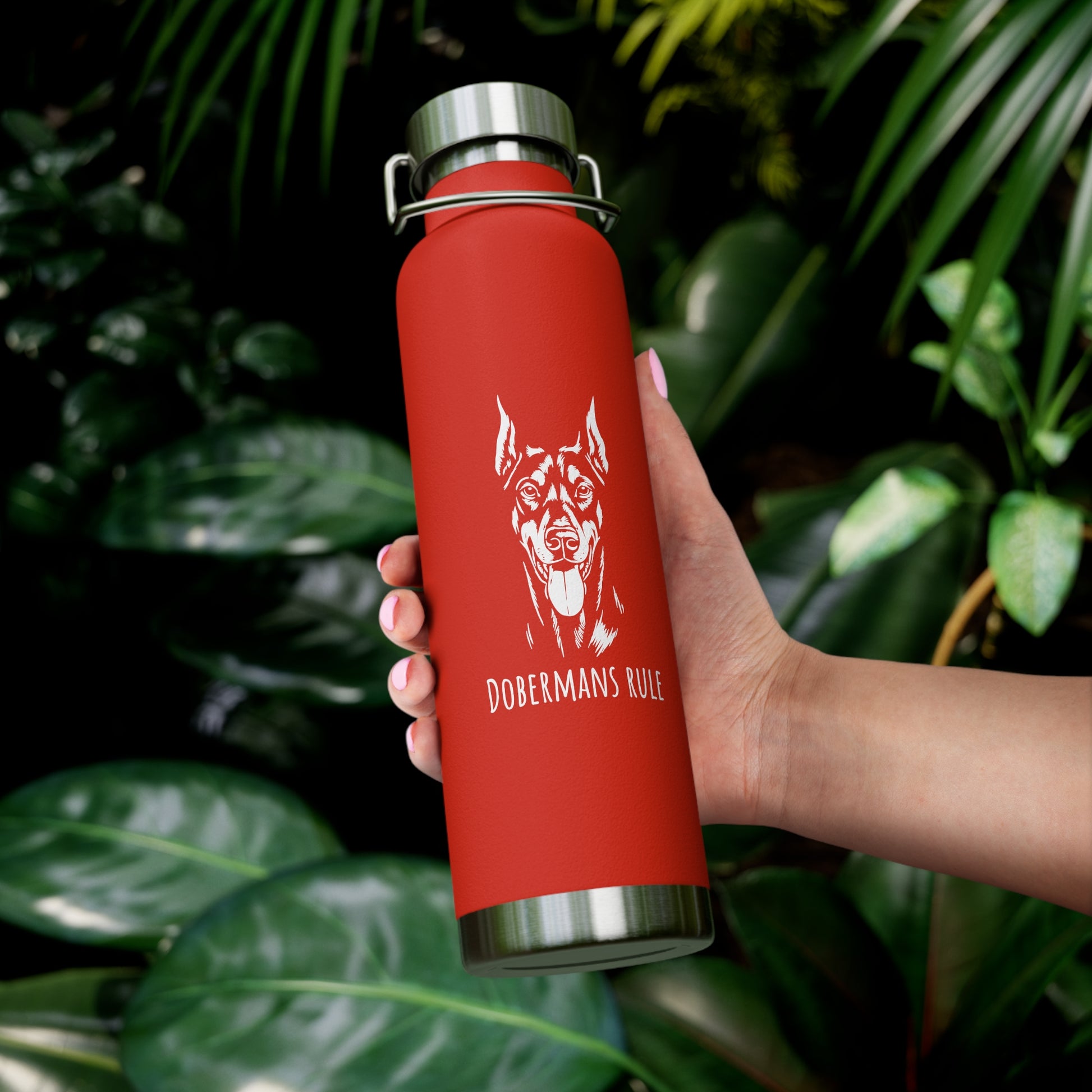 Red water bottle with a dog illustration and 'Dobermans Rule' text held by a hand against a leafy background