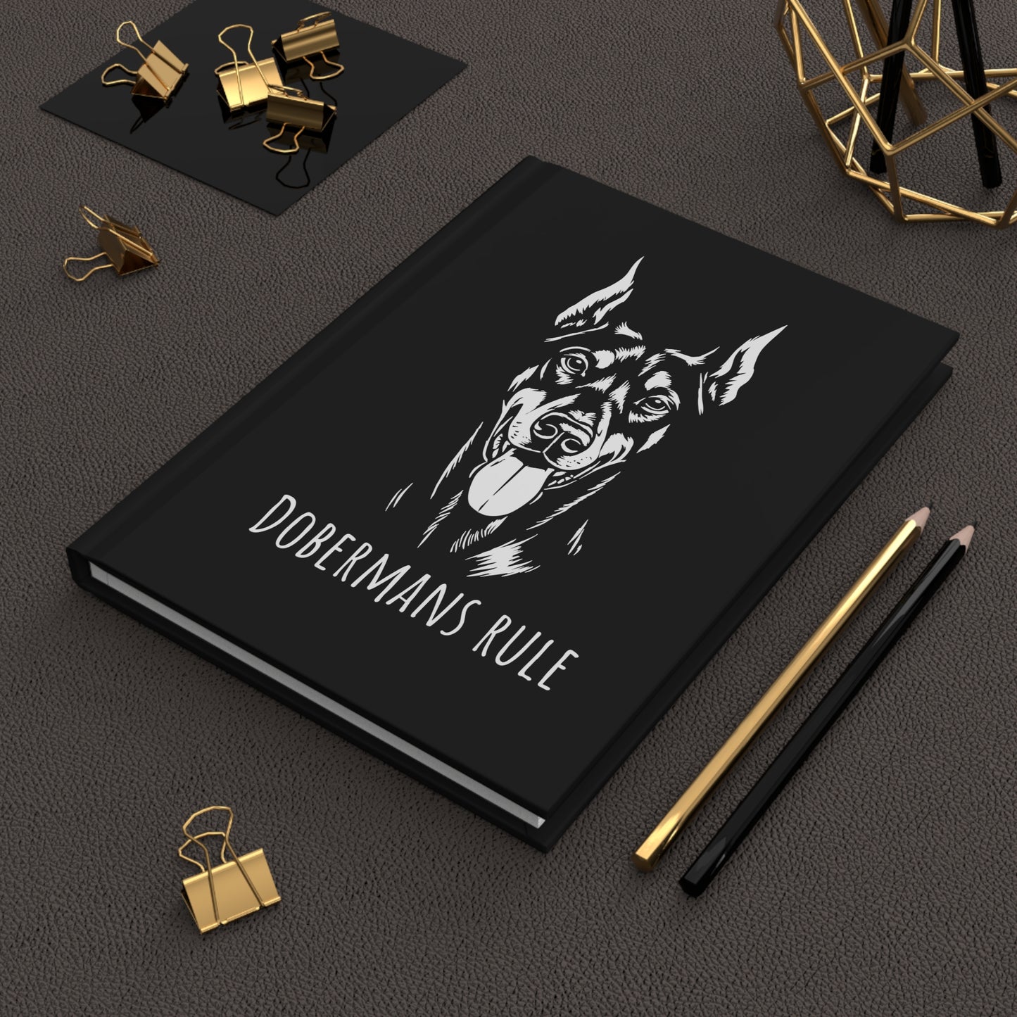 Black notebook with doberman illustration and 'Dobermans Rule' text on a dark surface with office supplies.
