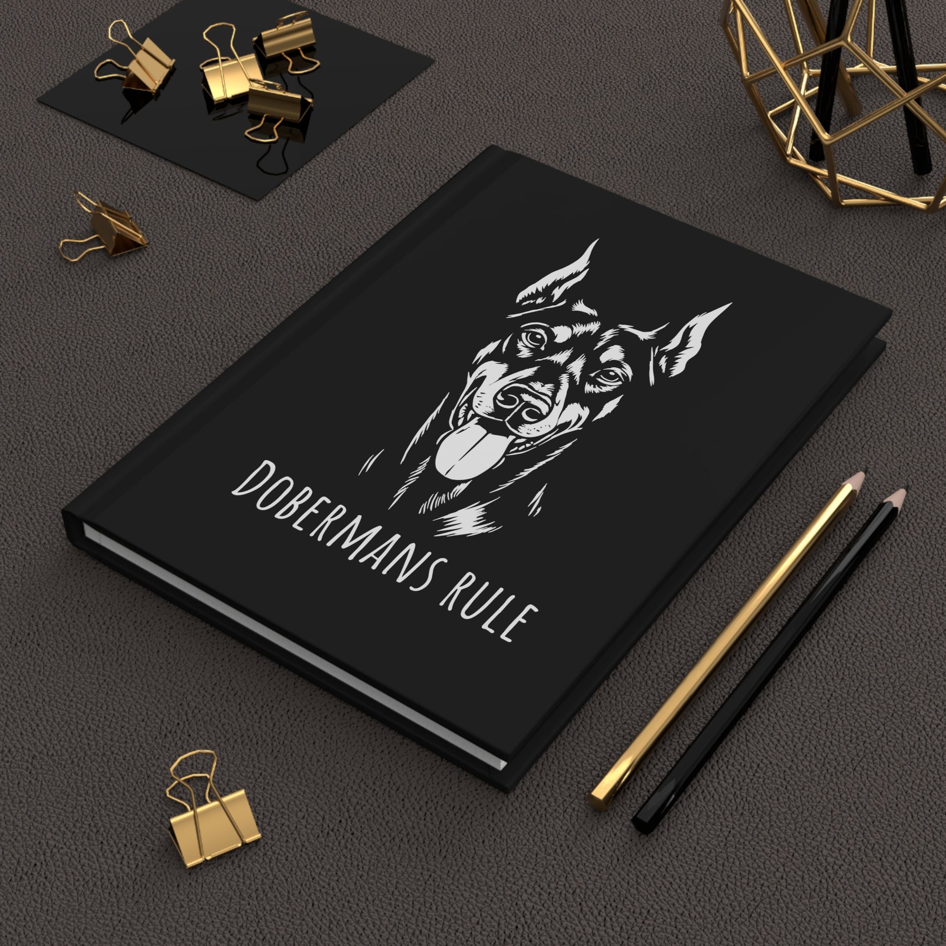 Black notebook with doberman illustration and 'Dobermans Rule' text on a dark surface with office supplies.