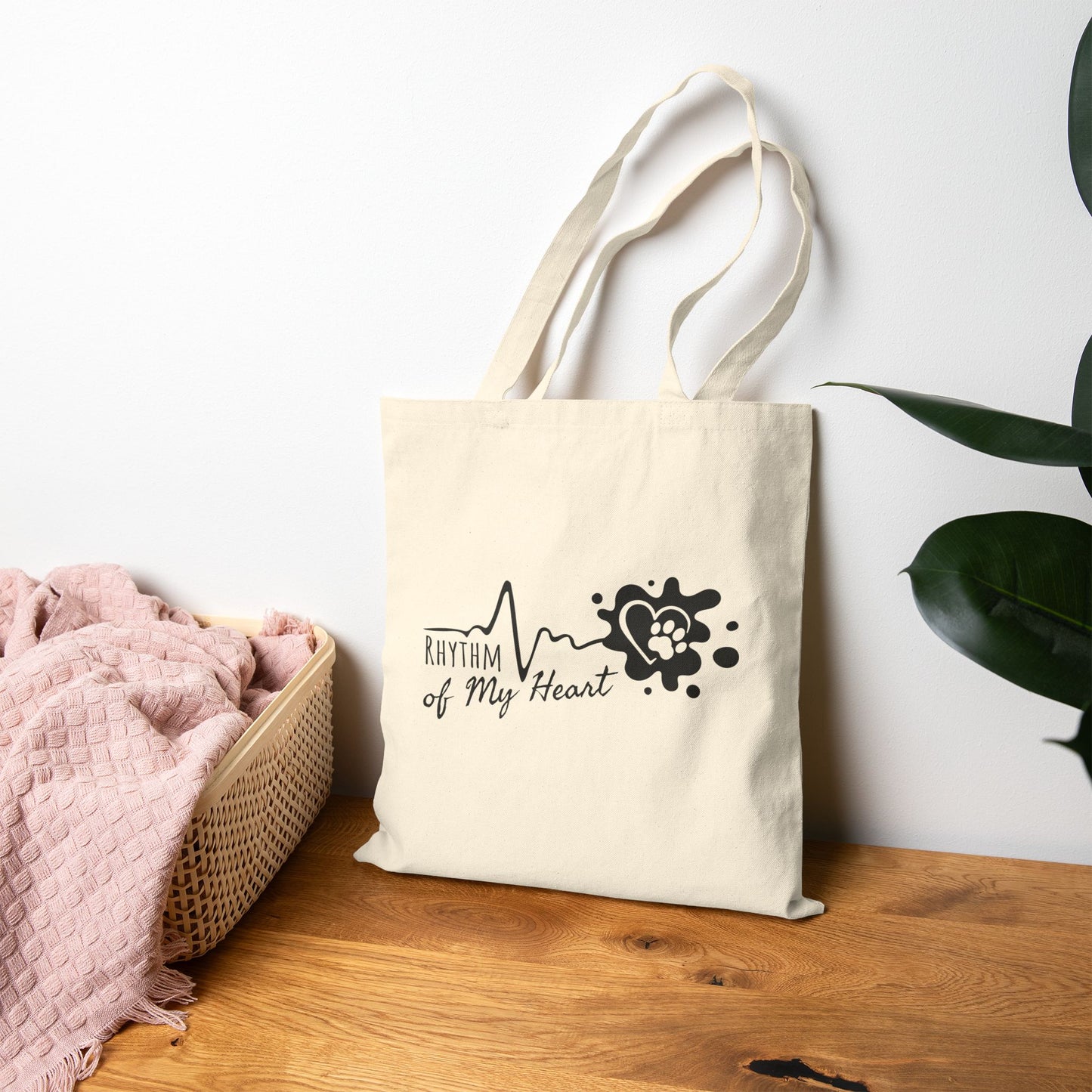 Beige tote bag with 'Rhythm of My Heart' design on a wooden surface with a white wall and plant in the background.