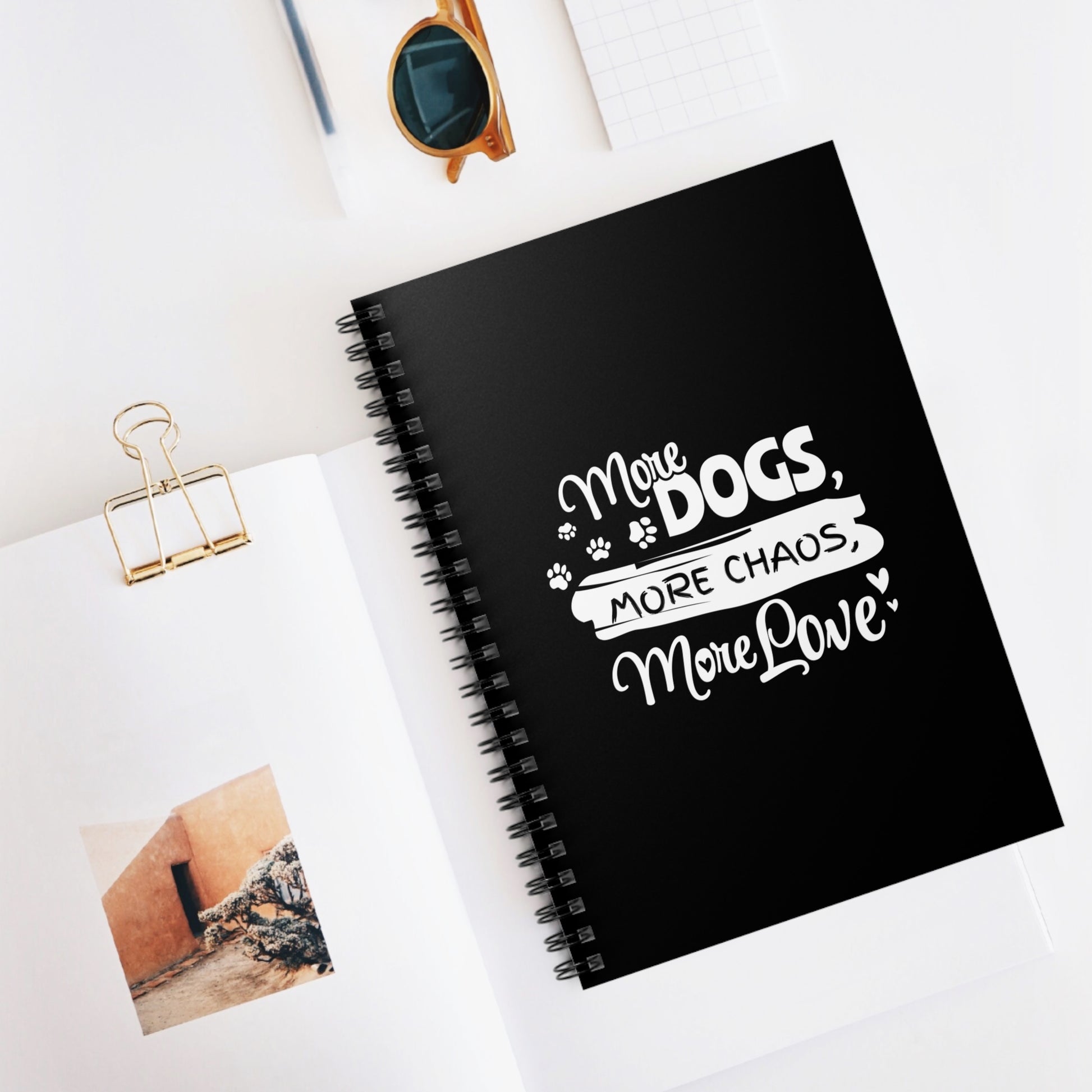 Black notebook with 'More Dogs, More Chaos, More Love' text on a white surface with photos and sunglasses.