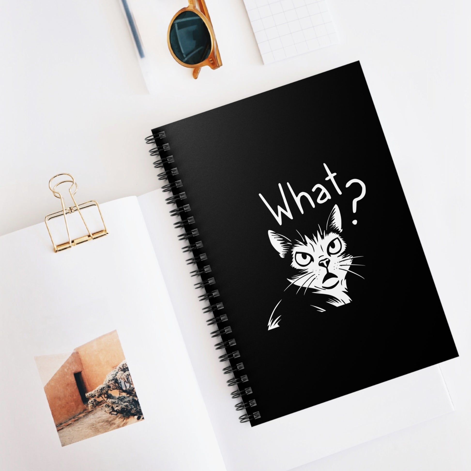 Black notebook with a cat illustration and 'What?' text on a white surface with sunglasses and a photo clip.