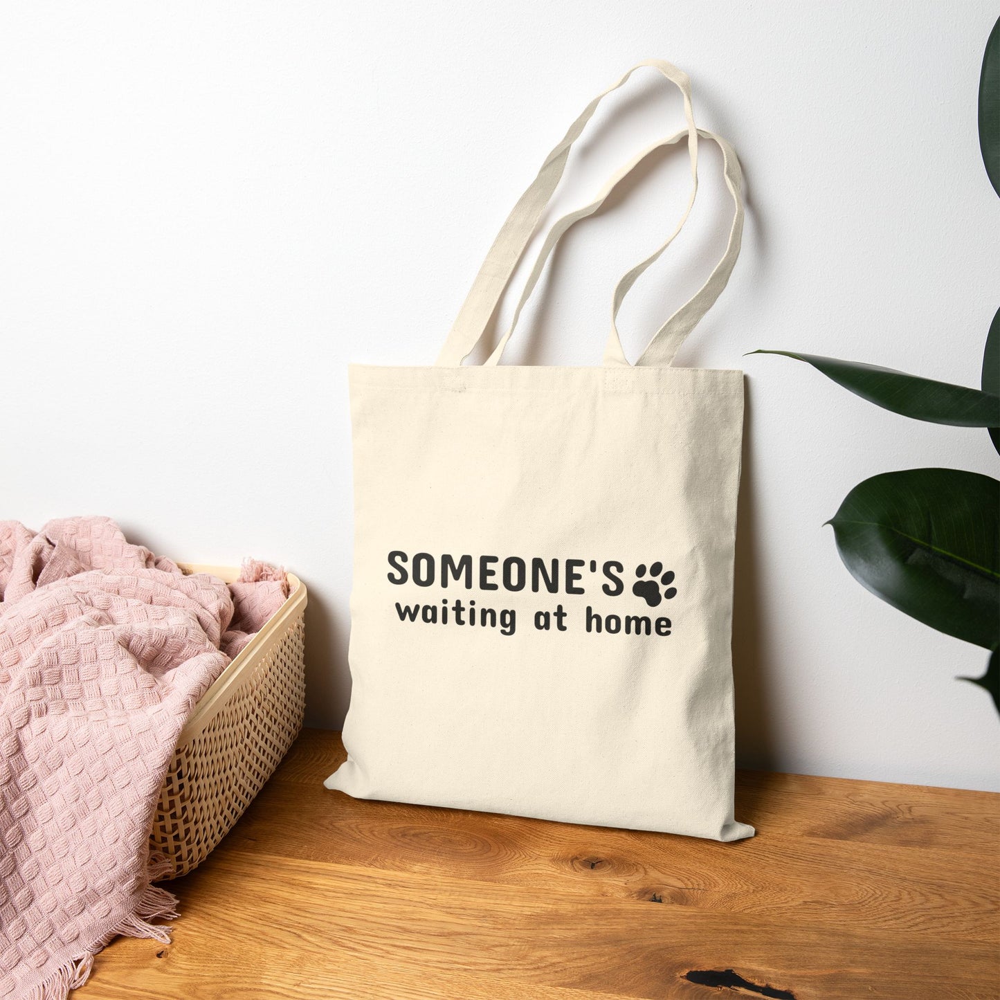 Beige tote bag with 'SOMEONE'S waiting at home' text on a wooden surface with a plant in the background